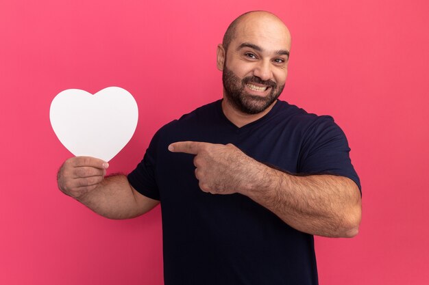 Hombre barbudo en camiseta con corazón de cartón apuntando con el dedo índice sonriendo con cara feliz de pie sobre la pared rosa