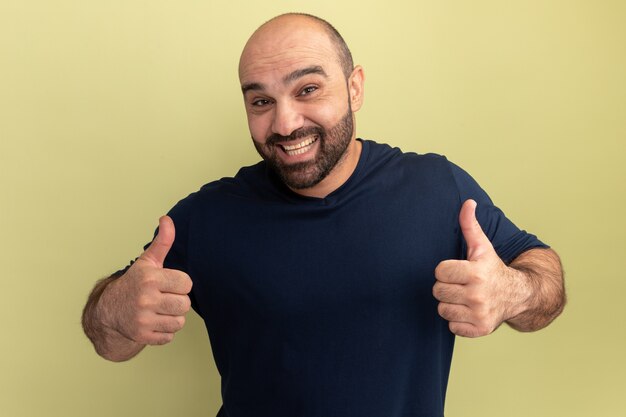 Hombre barbudo en camiseta negra feliz y emocionado mostrando los pulgares para arriba de pie sobre la pared verde