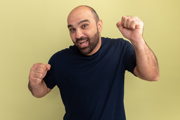 Hombre barbudo en camiseta negra loco feliz y emocionado gritando apretando los puños de pie sobre la pared verde