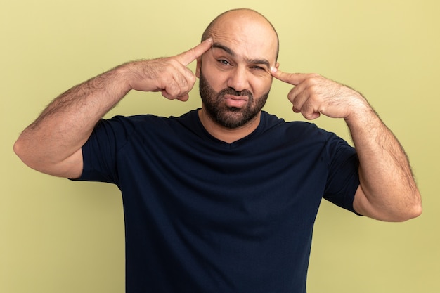 Foto gratuita hombre barbudo en camiseta negra molesto y cansado señalando con los dedos índices en las sienes de pie sobre la pared verde