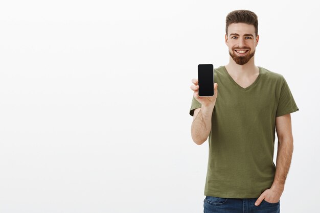 Hombre barbudo carismático, guapo, confiado y complacido, mostrando la pantalla del teléfono inteligente y sonriendo felizmente
