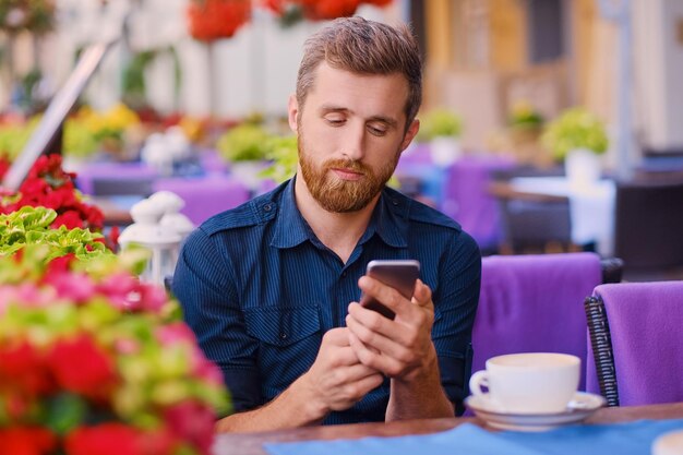 Hombre barbudo casual bebe café y usa un teléfono inteligente en un café