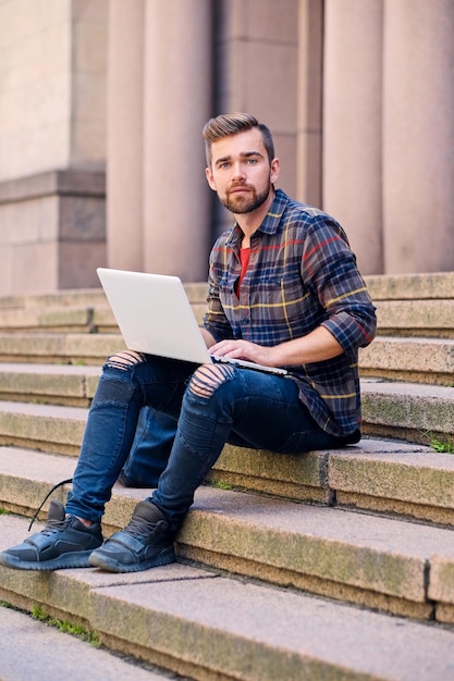 Un hombre barbudo casual vestido con jeans y camisa polar se sienta en un escalón y usa una computadora portátil.