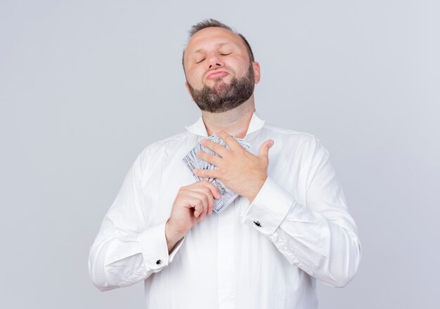 Hombre barbudo complacido vestido con camisa blanca sosteniendo efectivo mirando confiado de pie sobre la pared blanca