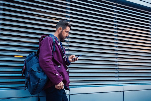 Hombre barbudo confiado sosteniendo un teléfono móvil mientras está de pie cerca de la pared de la calle en una soleada noche de verano.