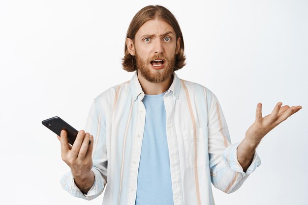 Hombre barbudo confundido que parece desconcertado sosteniendo el teléfono móvil y encogiéndose de hombros sin idea no puede entender algo en línea de pie sobre fondo blanco