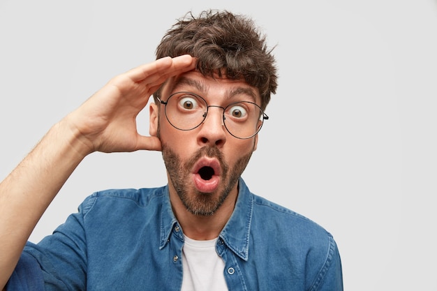 Foto gratuita el hombre barbudo conmocionado y emocionado mira fijamente con ojos saltones, mantiene la mano en la cabeza, expresa incredulidad, tiene una reacción silenciosa, aislado sobre una pared blanca.