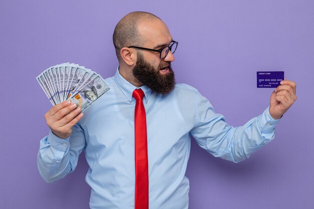 Hombre barbudo con corbata roja y camisa azul con gafas con dinero en efectivo y tarjeta de crédito mirándolo con una sonrisa en la cara feliz y positiva de pie sobre fondo púrpura