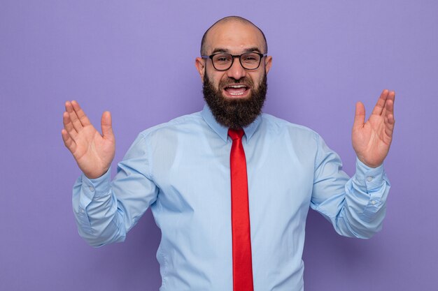 Hombre barbudo con corbata roja y camisa azul con gafas mirando feliz y emocionado gritando levantando las manos