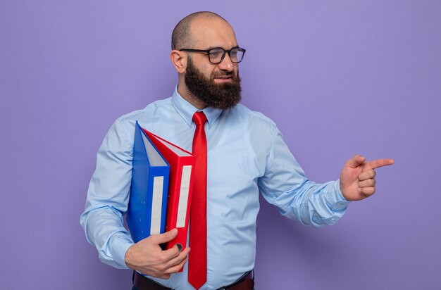 Hombre barbudo con corbata roja y camisa azul con gafas sosteniendo carpetas de oficina mirando a un lado con una sonrisa en la cara apuntando con el dedo índice hacia un lado