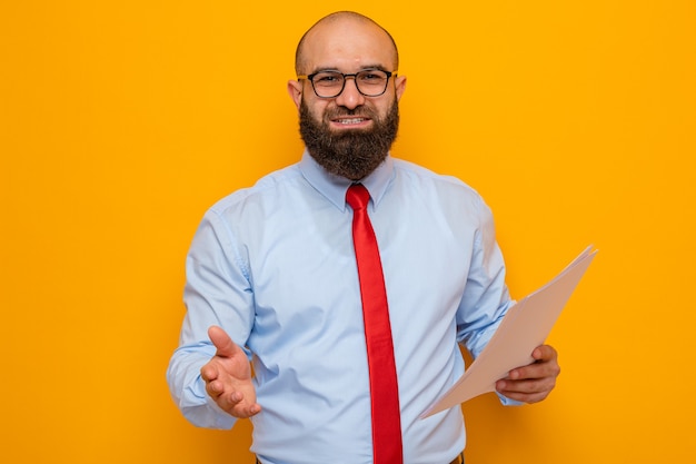 Hombre barbudo con corbata roja y camisa azul con gafas sosteniendo documentos mirando ofreciendo saludo de mano