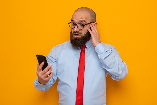 Hombre barbudo con corbata roja y camisa azul con gafas sosteniendo smartphone mirándolo asombrado y sorprendido de pie sobre fondo naranja