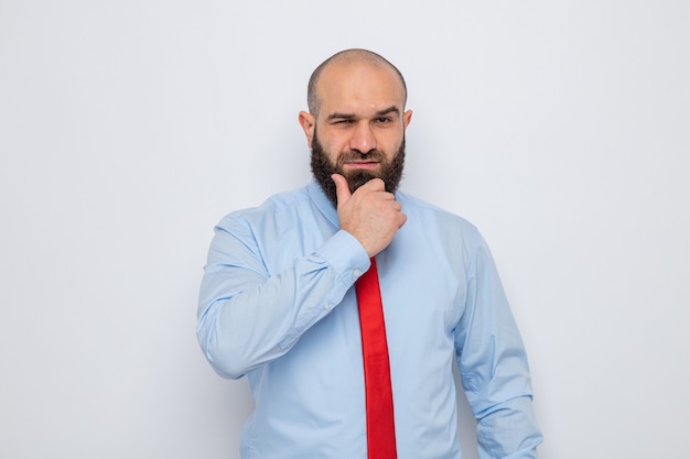 Hombre barbudo con corbata roja y camisa azul mirando con han en su barbilla con expresión pensativa pensando