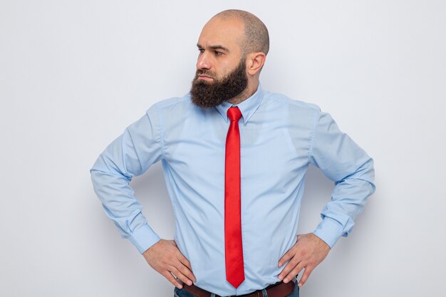 Hombre barbudo con corbata roja y camisa azul mirando a un lado con cara seria con las manos en la cadera