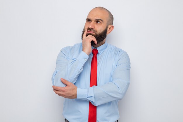 Hombre barbudo con corbata roja y camisa azul mirando a un lado con expresión pensativa con la mano en la barbilla de pie sobre fondo blanco.