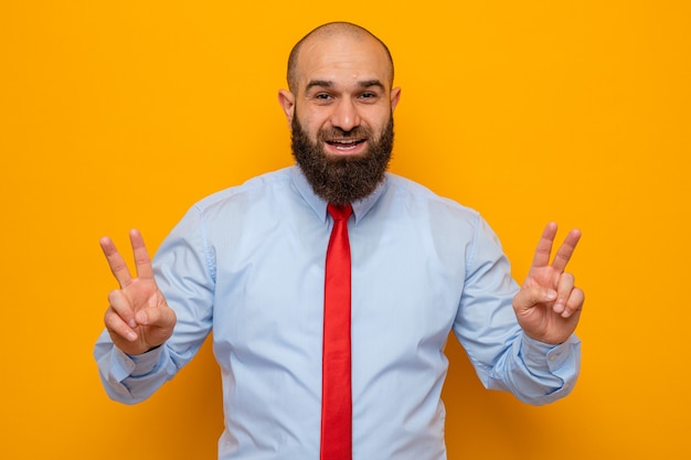 Hombre barbudo con corbata roja y camisa mirando feliz y alegre sonriendo ampliamente mostrando v-sign