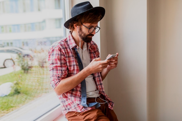 Hombre barbudo elegante en camisa a cuadros brillante que instala la nueva aplicación móvil en el dispositivo de teléfono inteligente y escucha música. Estilo hipster.