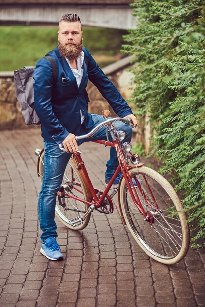 Hombre barbudo con un elegante corte de pelo vestido con ropa informal con una mochila, sentado en una bicicleta retro en un parque de la ciudad.