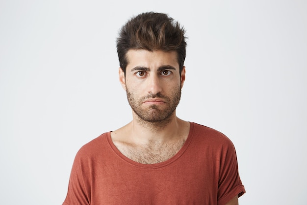 Hombre barbudo elegante que disgustó la mirada de pie en tela casual contra la pared blanca. Joven tiene algunos problemas para mirar con insatisfacción y descontento