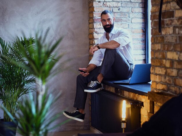 Hombre barbudo elegante trabaja con una computadora portátil en una habitación con interior de loft.