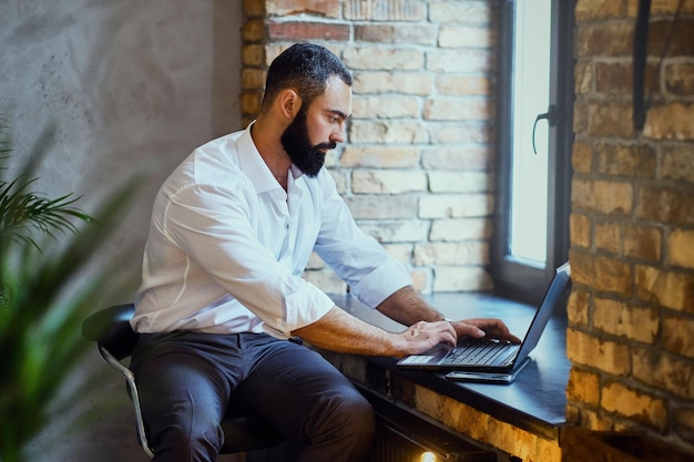 Hombre barbudo elegante trabaja con una computadora portátil en una habitación con interior de loft.