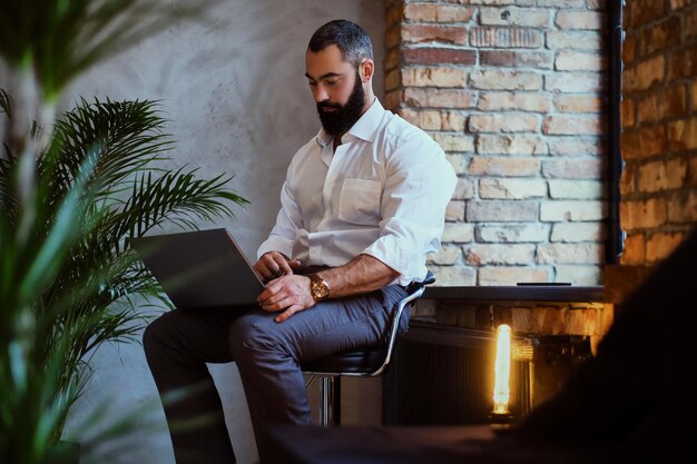 Hombre barbudo elegante trabaja con una computadora portátil en una habitación con interior de loft.