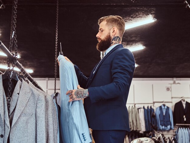 Un hombre barbudo elegantemente vestido con tatuajes en las manos y el cuello elige un nuevo traje en la tienda de ropa masculina.