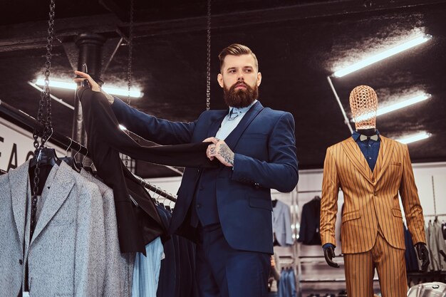 Un hombre barbudo elegantemente vestido con tatuajes en las manos y el cuello elige un traje nuevo en una tienda de ropa masculina.