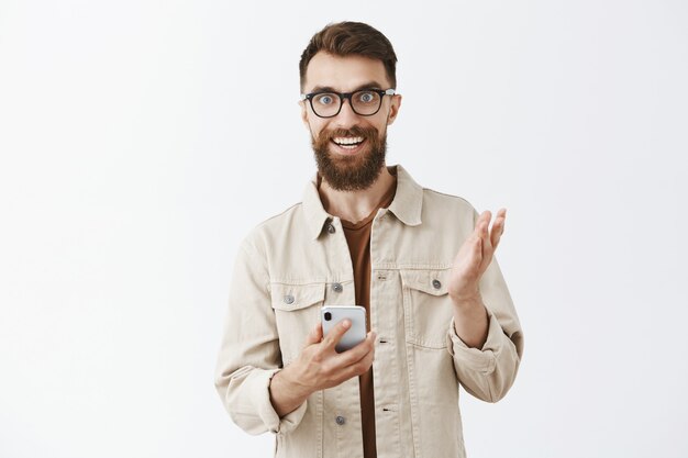 Hombre barbudo emocionado con gafas posando contra la pared blanca