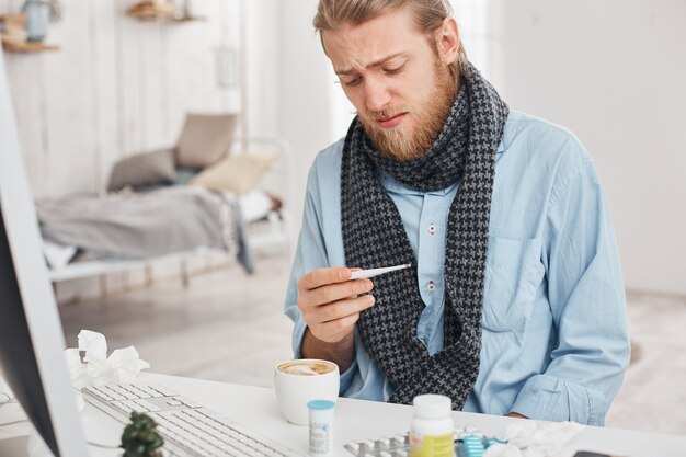 El hombre barbudo enfermo o enfermo usa un termómetro para medir la temperatura de su cuerpo. El hombre rubio mira desesperadamente el termómetro, sufre de un fuerte resfriado, rodeado de medicamentos en su lugar de trabajo.