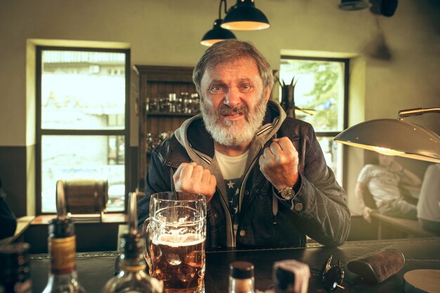 Hombre barbudo enojado bebiendo alcohol en un pub y viendo un programa deportivo en la televisión.