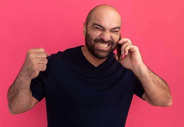 Hombre barbudo enojado en camiseta azul marino que parece molesto mientras habla por teléfono móvil apretando el puño parado sobre la pared rosa