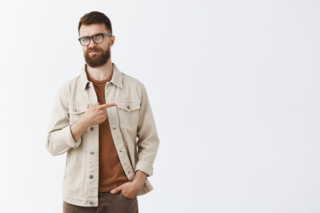 Hombre barbudo escéptico y decepcionado con gafas posando contra la pared blanca