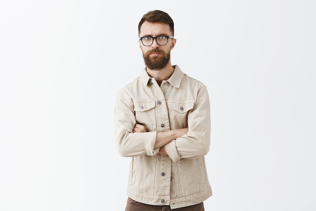 Hombre barbudo escéptico en gafas posando contra la pared blanca