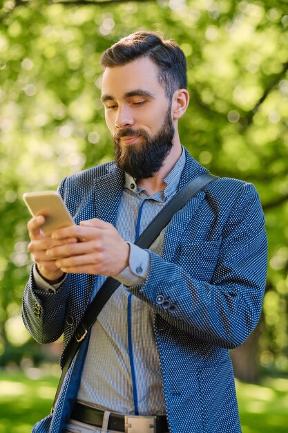 Hombre barbudo con estilo en una chaqueta azul usando un teléfono inteligente en un parque.