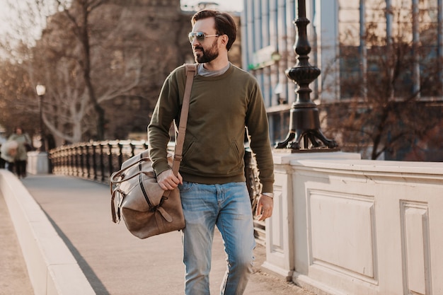 Hombre barbudo con estilo guapo caminando en la calle de la ciudad con bolsa de viaje de cuero con sudadera y gafas de sol, tendencia de estilo urbano, día soleado, confiado y sonriente