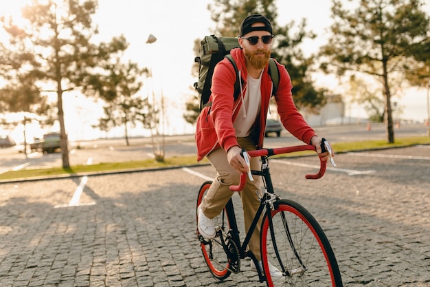 Hombre barbudo estilo hipster guapo con capucha roja y gafas de sol montando solo con mochila en bicicleta mochilero de viajero de estilo de vida activo saludable