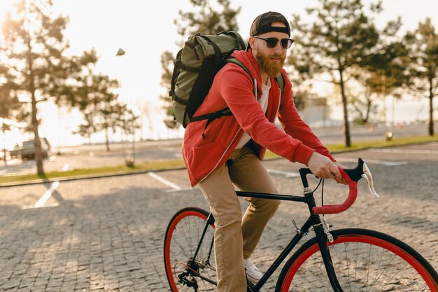 Hombre barbudo estilo hipster guapo con capucha roja y gafas de sol montando solo con mochila en bicicleta mochilero de viajero de estilo de vida activo saludable