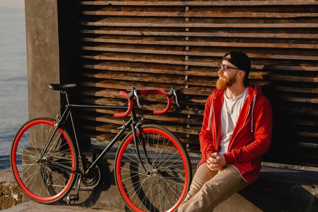 Hombre barbudo de estilo hipster guapo con capucha roja sentado relajándose solo con mochila y bicicleta en el amanecer de la mañana junto al mar mochilero de viajero de estilo de vida activo saludable