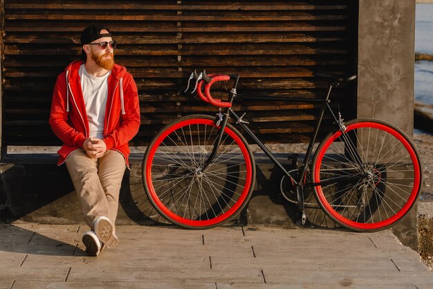 Hombre barbudo de estilo hipster guapo con capucha roja sentado relajándose solo con mochila y bicicleta en el amanecer de la mañana junto al mar mochilero de viajero de estilo de vida activo saludable