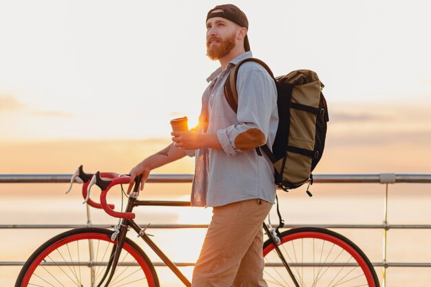 Hombre barbudo de estilo hipster guapo con mochila con camisa de mezclilla y gorra con bicicleta en el amanecer de la mañana junto al mar tomando café, mochilero viajero de estilo de vida activo y saludable