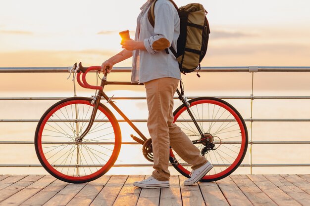 Hombre barbudo de estilo hipster guapo con mochila con camisa de mezclilla y gorra con bicicleta en el amanecer de la mañana junto al mar tomando café, mochilero viajero de estilo de vida activo y saludable