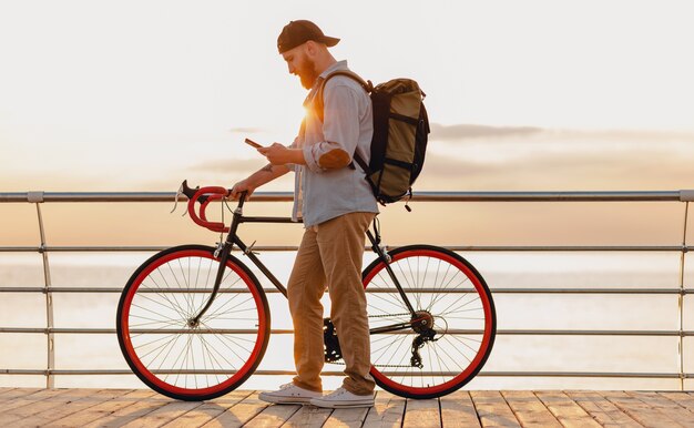 Hombre barbudo de estilo hipster guapo con mochila con camisa de mezclilla y gorra viajando con bicicleta en el amanecer de la mañana junto al mar tomando café, mochilero viajero de estilo de vida activo saludable