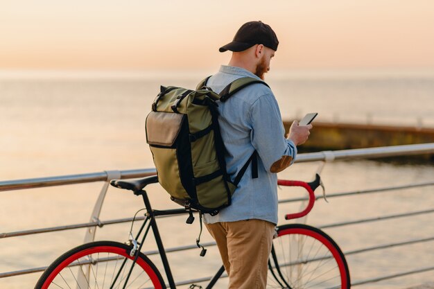 Hombre barbudo de estilo hipster guapo usando teléfono viajando con mochila y bicicleta en el amanecer de la mañana junto al mar, mochilero viajero de estilo de vida activo saludable