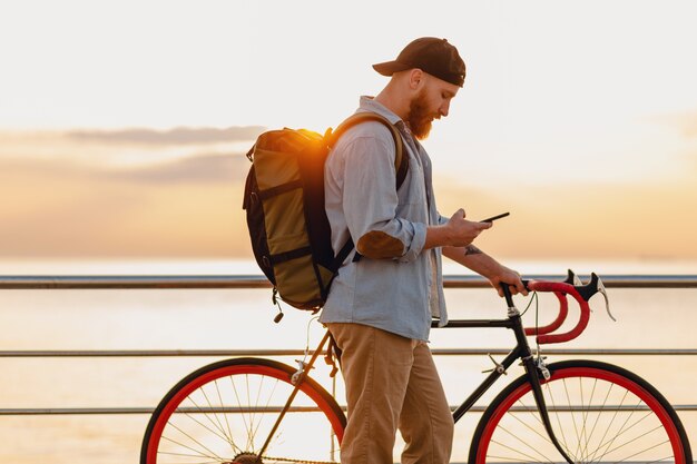 Hombre barbudo de estilo hipster guapo usando teléfono viajando con mochila y bicicleta en el amanecer de la mañana junto al mar, mochilero viajero de estilo de vida activo saludable