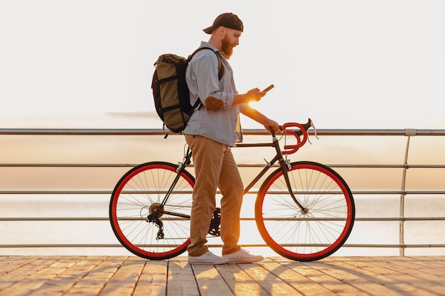 Hombre barbudo de estilo hipster guapo viajando con mochila en bicicleta usando el teléfono en el amanecer por la mañana junto al mar, estilo de vida activo y saludable