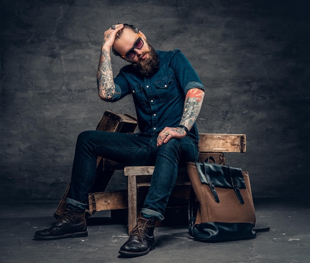 Hombre barbudo con estilo en jeans y gafas de sol se sienta en una caja de madera sobre fondo gris.