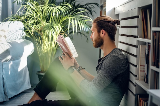 Hombre barbudo con estilo leyendo un libro en una habitación con plantas verdes.
