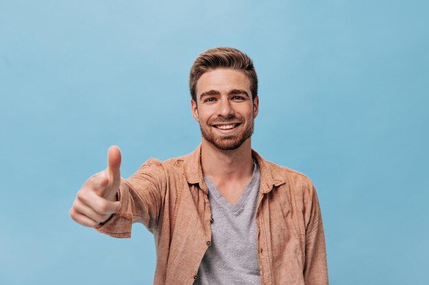 Hombre barbudo feliz con corte de pelo corto en camisa beige y camiseta fresca apuntando a la cámara y sonriendo en un contexto aislado