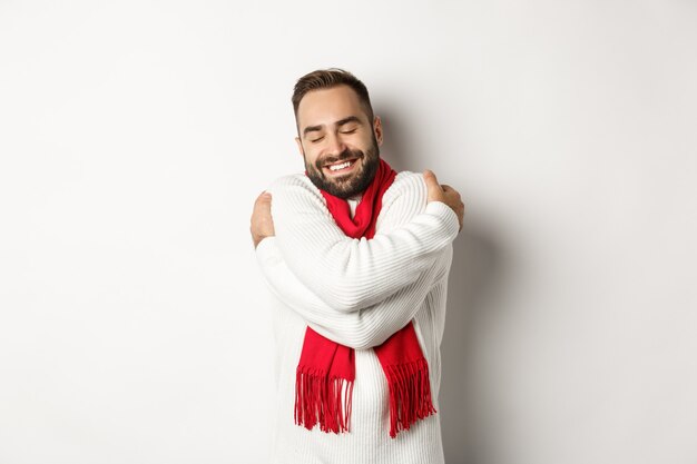 Hombre barbudo feliz disfrutando de un cómodo suéter de invierno, sonriendo con calidez, abrazándose a sí mismo, de pie sobre fondo blanco.